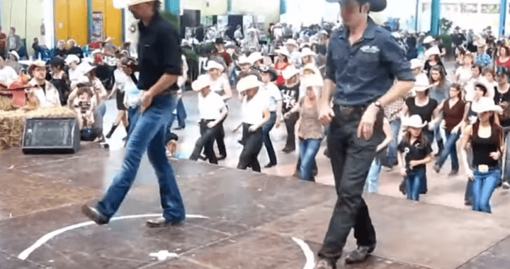 Two Men In Cowboy Outfits Lead Crowd In A Line Dance